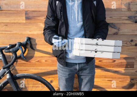 La persona di consegna in piedi accanto ad una bicicletta tiene scatole di pizza e un telefono. Lavoro come corriere, professione di messaggero di bici, concetto di lavoro part-time Foto Stock