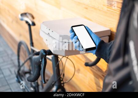 La persona di consegna in piedi accanto ad una bicicletta con le scatole della pizza tiene un telefono con lo schermo del whote. Lavoro come un corriere, professione del messaggero della bici, tempo parziale WO Foto Stock