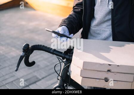 La persona di consegna in piedi accanto ad una bicicletta tiene scatole di pizza e un telefono. Lavoro come corriere, professione di messaggero di bici, concetto di lavoro part-time Foto Stock