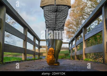 Basso angolo, vista posteriore della donna britannica in stivali wellington / wellies, isolati all'aperto a piedi in inverno sole che attraversa un ponte di legno. Foto Stock
