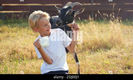 Un ragazzo beve latte di capra da una tazza accanto alla sua capra. Foto Stock