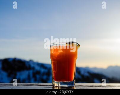 Bicchiere illuminato contenente cocktail arancione con zeppa di lime contro le montagne sfocate. Foto Stock