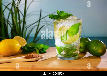 Primo piano di un cocktail mojito con limoni freschi, lime, foglie di menta e un cucchiaio di legno con zucchero di canna su una tavola di legno. Ingredienti freschi. Foto Stock