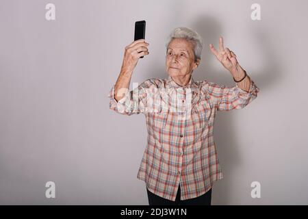 Vecchia donna che fa un selfie con un telefono intelligente e felice e il simbolo della pace o della vittoria in mano. Concetto tecnologico. Concetto attivo di vecchiaia. GRA Foto Stock