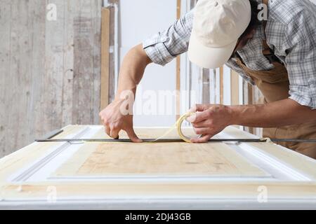 Falegname maschio che lavora il legno in officina di carpenteria, mettendo nastro di mascheratura di carta sulla porta di legno seguendo la linea di righello quadrato di metallo, indossando ove Foto Stock