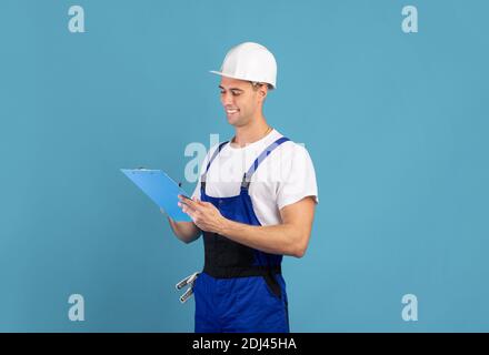 Sorridente appaltatore di riparazione maschile in tute e note di scrittura hardhat negli appunti Foto Stock