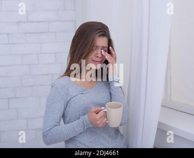 Triste giovane donna con tazza di caffè piangendo vicino alla finestra, strofinando le sue lacrime a casa Foto Stock