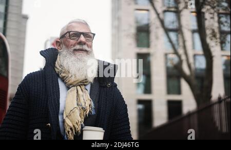 Hipster uomo d'affari senior che beve caffè mentre si cammina in ufficio Con la città sullo sfondo - Focus on Face Foto Stock