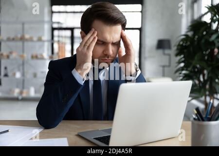 Stanco stressato uomo d'affari che tocca i templi, seduto alla scrivania con il computer portatile Foto Stock