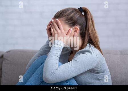 Vista laterale della giovane donna con depressione che copre il viso e piangendo sul divano a casa Foto Stock