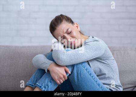 Giovane donna depressa seduta sul divano con gli occhi chiusi e abbracciando le ginocchia, sentendosi disperata e stressata Foto Stock