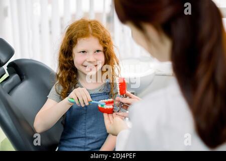 Vista posteriore della giovane donna caucasica dentista che tiene le mascelle artificiali, mentre la bambina piccola riccia spazzola i denti con spazzolino da denti e sorridendo al medico Foto Stock