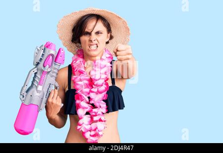 Bella giovane donna con capelli corti indossando bikini e hawaian lei tenere pistola acqua irritata e frustrata gridando con rabbia, urlando pazzo con Foto Stock