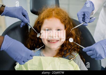 Piccola ragazza europea di charme con capelli ricci rossi, seduta in sedia dentale, sorridente e guardando la macchina fotografica durante il trattamento medico a dentale moderno Foto Stock