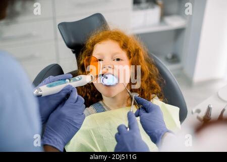 Vista ravvicinata della bambina che ha un trattamento dentale presso l'ufficio del dentista. Mani di due dentisti femminili che fanno il restauro del dente con riempimento Foto Stock