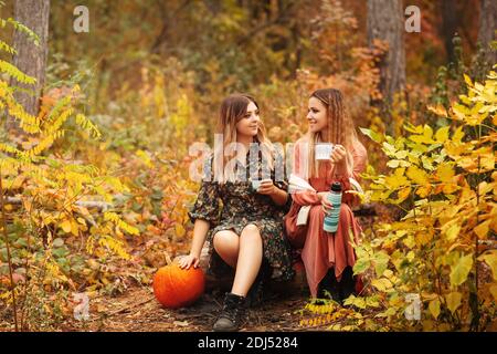 Giovani amiche in abiti eleganti che si guardano l'un l'altro e gustando una bevanda calda mentre si siedono sulle zucche in autunno foresta Foto Stock