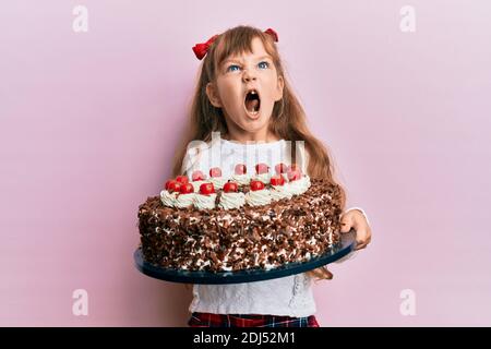 Piccolo caucasico ragazza capretto festeggiando il compleanno tenendo grande torta di cioccolato arrabbiato e pazzo urlando frustrato e furioso, gridando con rabbia. Rabbia un Foto Stock