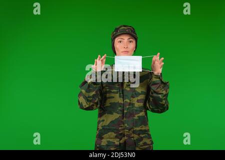 Donna in uniforme militare tiene una maschera medica di fronte con entrambe le mani. Il concetto di stereotipi di genere e pandemia. Foto Stock