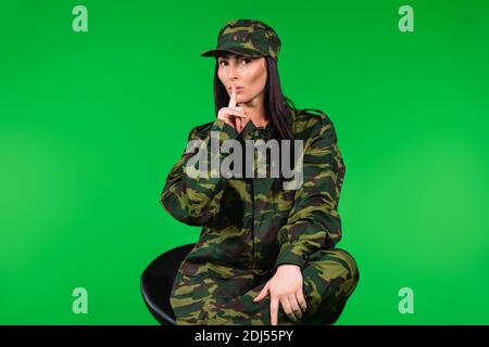 Giovane bella donna in uniforme militare in posa mentre si siede su una sedia, gesturando shh con dito indice. Stereotipi di genere. Foto Stock