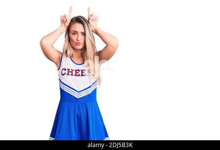 Giovane bella donna bionda che indossa uniforme cheerleader facendo gesto divertente con il dito sulla testa come corna toro Foto Stock