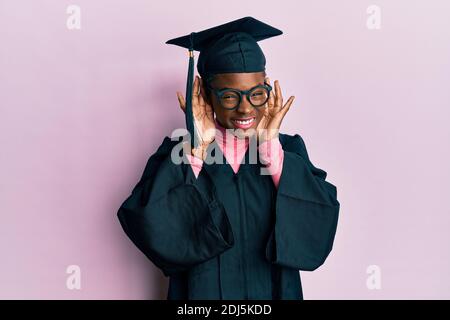 Giovane afroamericana ragazza con cappuccio di graduazione e accappatoio cerimonia cercando di sentire entrambe le mani sul gesto dell'orecchio, curiosa per pettegolezzi. Problemi di udito, de Foto Stock