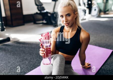 Giovane sportivo in abbigliamento sportivo si siede sul pavimento su un tappetino fitness, in palestra e beve acqua dopo l'allenamento, giocando sport Foto Stock