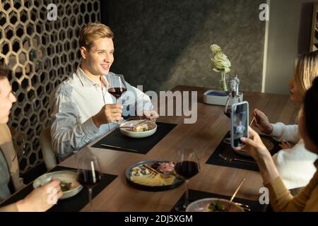 Felice giovane elegante maschio con bicchiere di vino rosso brindisi con cena mentre sedendo al tavolo servito in moderno ristorante tra amici Foto Stock