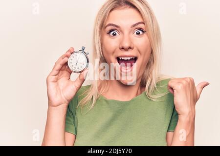 Giovane bella donna bionda che fa conto alla rovescia usando cronometro su bianco sfondo che punta il pollice verso il lato sorridendo felice con aprire la bocca Foto Stock