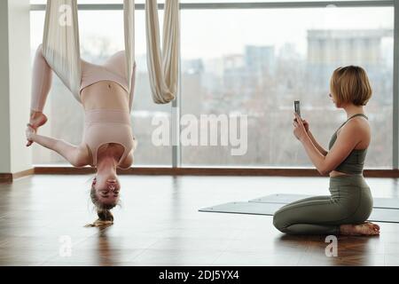 Giovane donna con smartphone che scatta foto di un'altra donna in forma in activewear appeso su amaca bianca durante lo yoga difficile della mosca esercizio Foto Stock