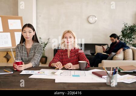 Giovane lavoratrice asiatica e la sua collega bionda matura avere caffè mentre si siede a tavola con molti documenti contro uomo d'affari Foto Stock