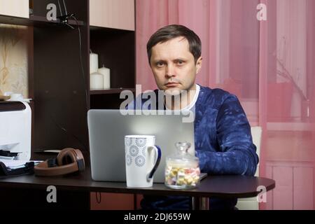 Un uomo in casa vestiti funziona a un computer portatile. Guarda nella fotocamera. Foto Stock