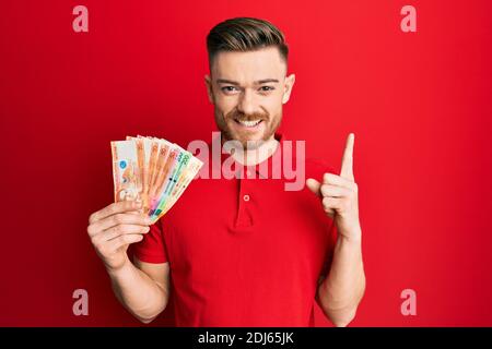 Giovane uomo rosso che tiene le banconote di peso filippino sorridendo con un'idea o una domanda che punta il dito con il volto felice, il numero uno Foto Stock