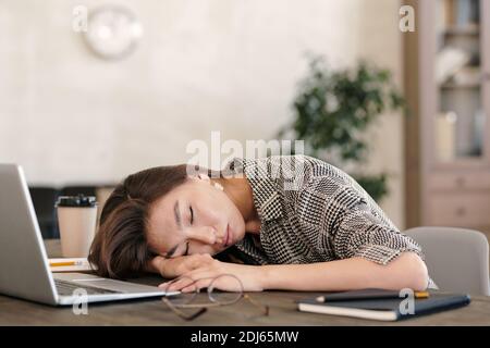 Giovane donna d'affari esausta in formalwear tenendo la testa a portata di mano mentre si annodano davanti al computer portatile dopo il lavoro in ufficio ambiente Foto Stock