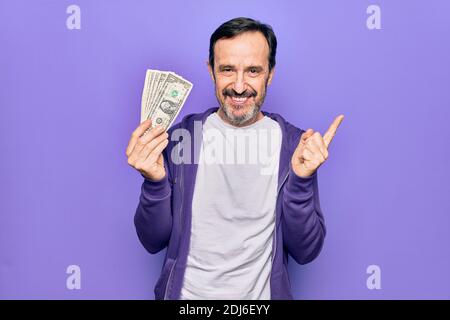 Uomo bello di mezza età che tiene mazzo di banconote di dollari sopra isolato sfondo viola sorridente felice puntando con mano e dito di lato Foto Stock