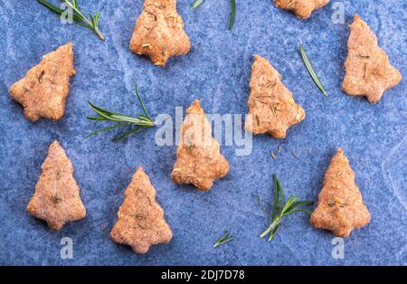 Biscotti fatti in casa a forma di albero di Natale cotti a base di grano integrale e formaggio, aromatizzati con rosmarino su una superficie di pietra blu Foto Stock