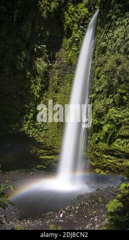 Un'esposizione verticale lunga ripresa di una cascata sottile con un bellissimo arcobaleno che scorre attraverso una scogliera mussosa Foto Stock