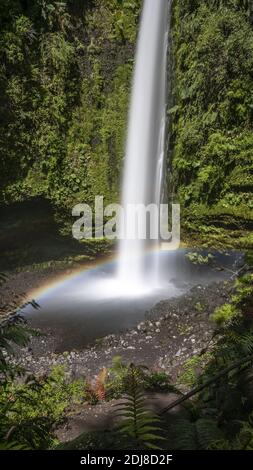 Un'esposizione verticale lunga ripresa di una cascata sottile con un bellissimo arcobaleno che scorre attraverso una scogliera mussosa Foto Stock