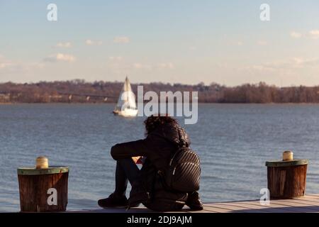 Una donna che indossa abiti scuri tra cui sneakers, cappotto invernale e zaino è seduta da sola su un molo che guarda al mare. Immagine per umore giù, premere Foto Stock