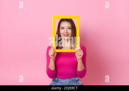 Primo piano ritratto di una giovane donna europea con un volto all'interno di una cornice vuota gialla, che tiene in mano, isolata su sfondo rosa Foto Stock