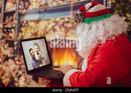 Babbo Natale sta guardando il portatile. Un uomo in un vestito di Natale al computer sullo sfondo delle luci di Capodanno. Il moderno mago. Natale h Foto Stock