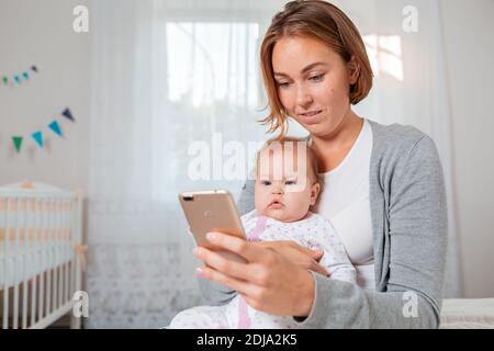 Ritratto di giovane madre tiene un bambino carino tra le braccia e gli mostra qualcosa sul suo smartphone. Concetto di intrattenimento online. Foto Stock