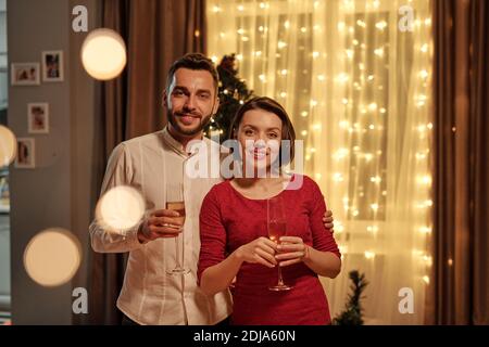 Ritratto di sorridente bell'uomo abbracciando bella ragazza e champagne flauti in un accogliente soggiorno con finestra decorata Foto Stock