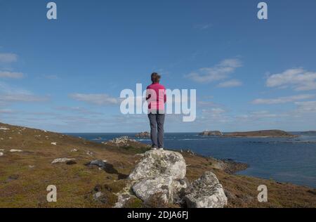 Femmina in piedi su una roccia di granito che domina l'Oceano Atlantico E Round Island Lighthouse sull'isola di Tresco in Le isole di Scilly Foto Stock