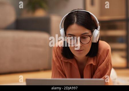 Ritratto di donna contemporanea che lavora da casa mentre si sdraiava sul pavimento indossando le cuffie e utilizzando il computer portatile, spazio per la copia Foto Stock