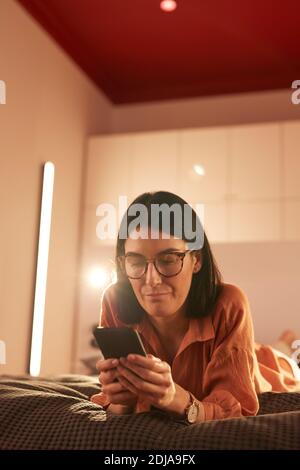 Ritratto verticale di una donna adulta contemporanea che usa lo smartphone mentre si sdraiava sul letto in interni minimal Foto Stock