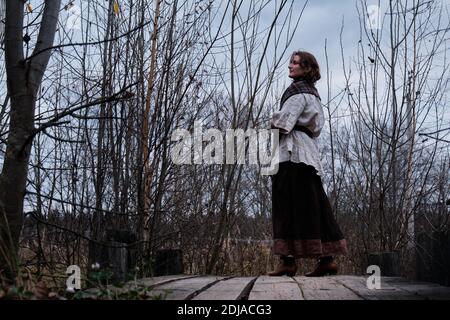 Una donna brunetta in abiti vintage è in piedi su una piattaforma vicino a una vecchia palude. Ritratto vicino a una palude con cespugli Foto Stock