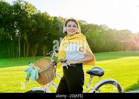 Bella donna di mezza età felice in cuffia con bicicletta nel parco Foto Stock