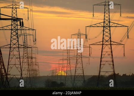 Sunset cielo & silhouette righe di energia elettrica linea tralicci & cavi che attraversano campagna paesaggio tramontare il sole scende sotto l'orizzonte Suffolk UK Foto Stock