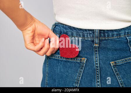 donna che tiene un piccolo cuore rosso frizzante mettendolo dentro o togliendo la tasca dei suoi jeans. Condividere e ricevere San Valentino, mantenere l'amore su di te Foto Stock