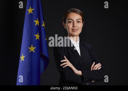 Ritratto professionale in vita della giovane dirigente femminile guardando la fotocamera e sorridendo mentre si sta in piedi con le braccia incrociate contro la bandiera dell'UE in background, Foto Stock